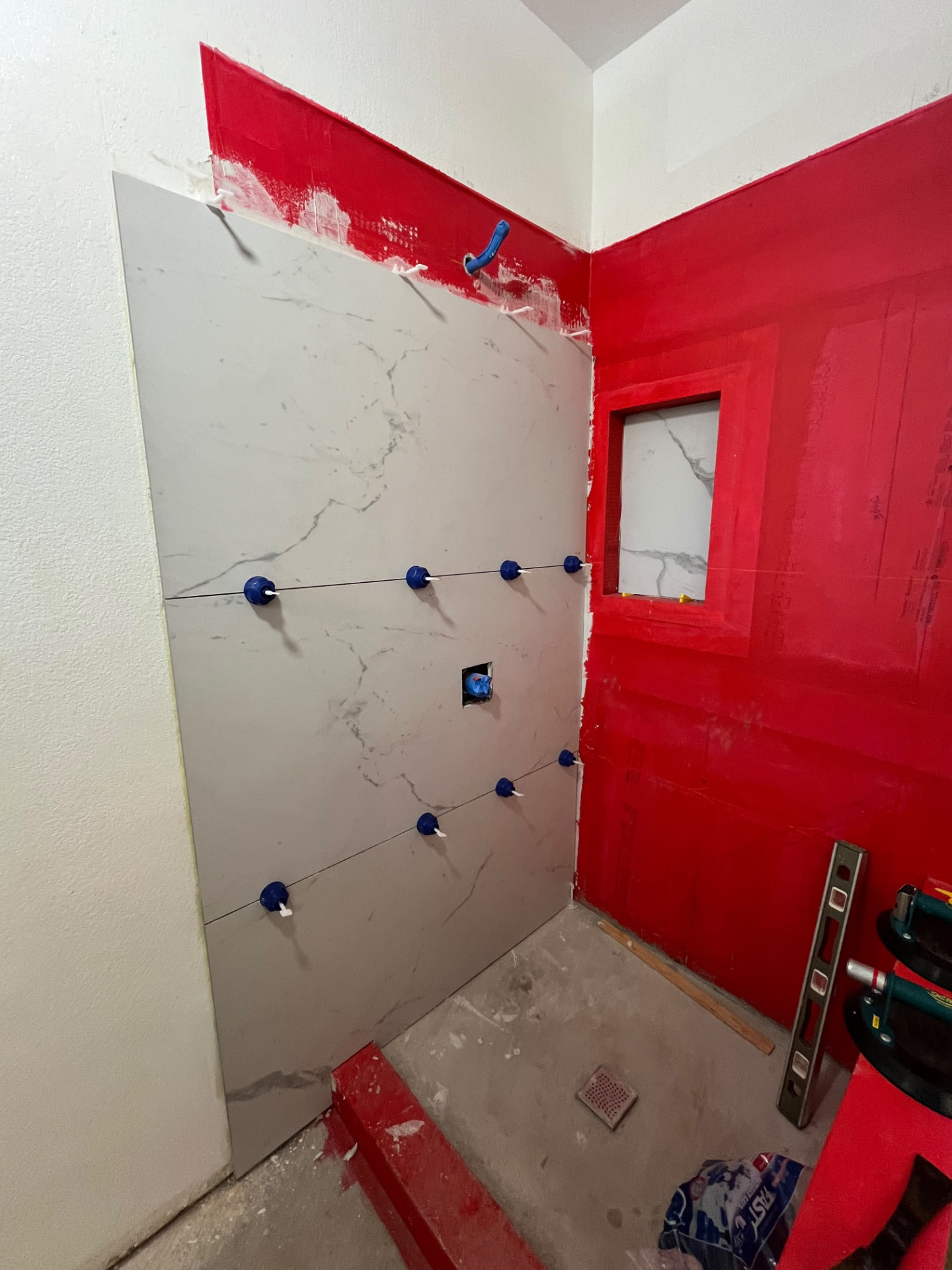 Large-format marble-look shower tile set with leveling clips over red waterproofing